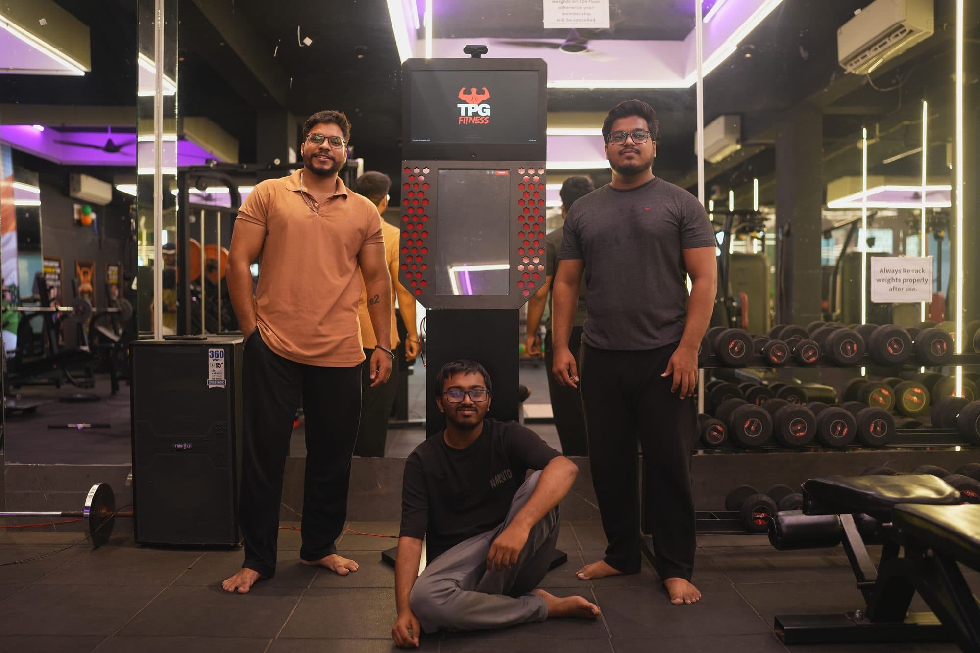Three TPG Fitness team members standing on a gym floor beside the kiosk, one crouched in front