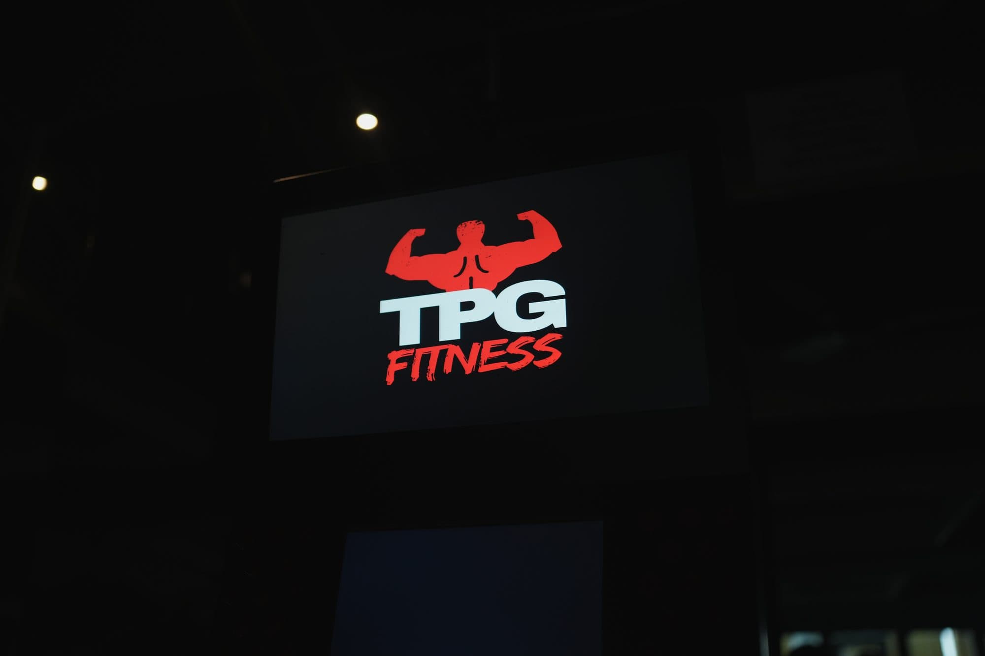 The TPG Fitness logo glowing on the kiosk's overhead screen in a dark gym