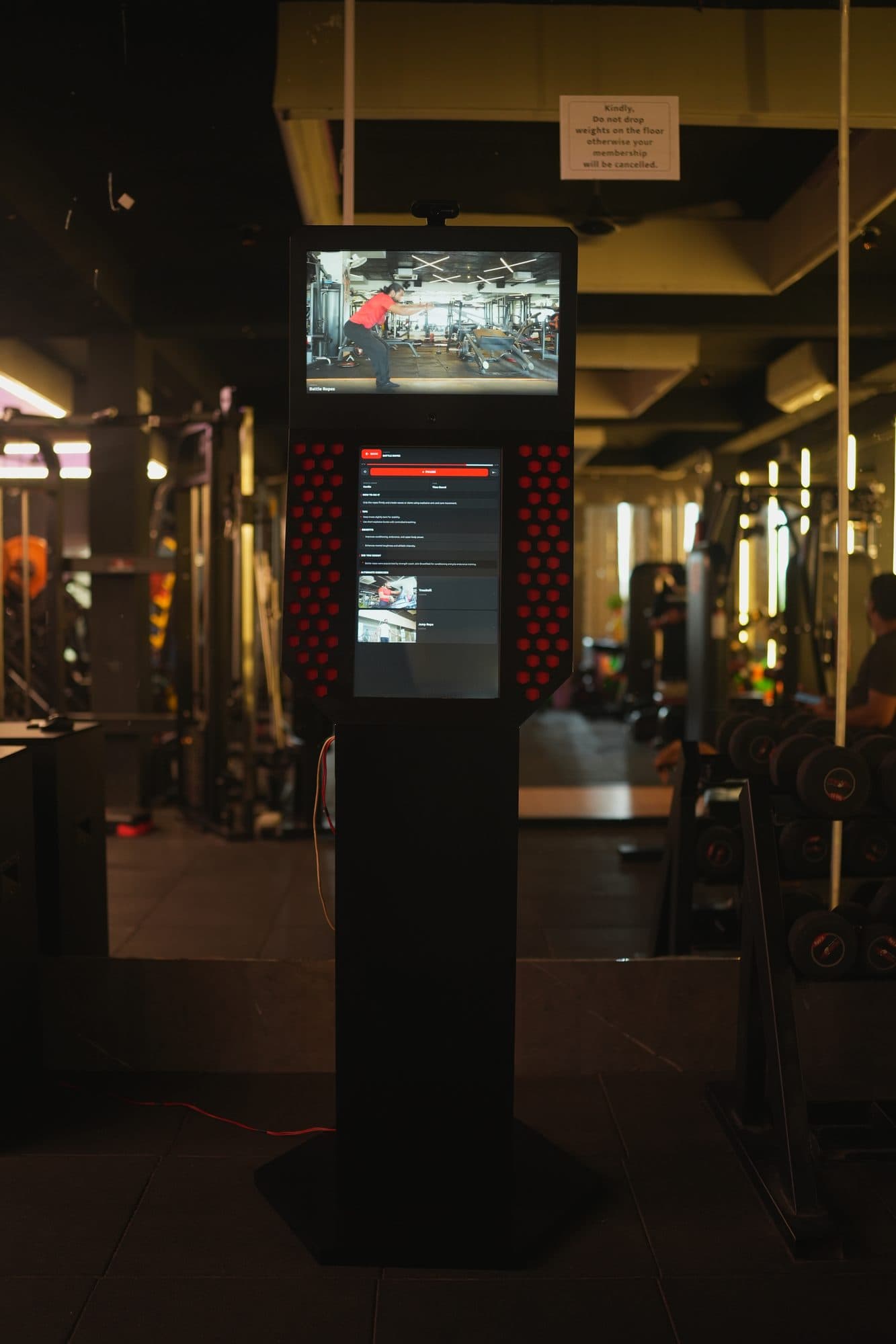 The kiosk installed on a gym floor with its screens on, surrounded by free weights