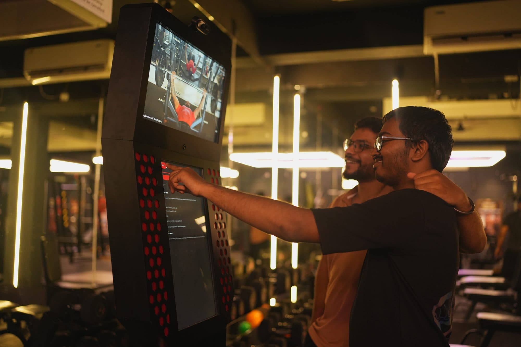 Two gym members close together at the kiosk, one selecting an exercise