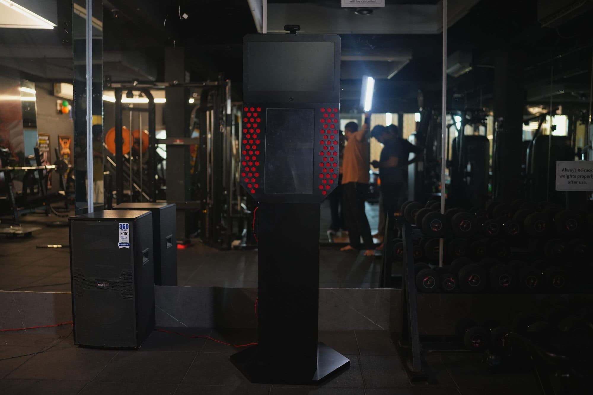 The kiosk on the gym floor at low light with equipment visible behind it